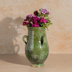 Op de afbeelding: Een groene keramische vaas met twee handvatten, gevuld met een boeket van levendige roze en paarse bloemen. De vaas heeft een rustieke, verouderde uitstraling, met zichtbare textuur en variaties in het groene glazuur.
