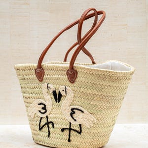 May include: Woven straw tote bag with brown leather handles. The bag features an embroidered design of two flamingos in cream and black. The handles are attached with brown leather patches. The interior of the bag is lined with white fabric.