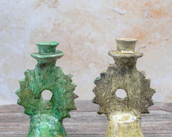 Puede incluir: Dos candeleros de cerámica. Uno es verde, el otro es marrón. Ambos tienen forma de sombrero de copa, una abertura circular y un diseño de sol. Están colocados sobre una superficie de madera.
