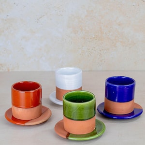Può includere: Quattro tazze da espresso e piattini in ceramica. Le tazze sono cilindriche con una base in terracotta e una parte superiore colorata in arancione, bianco, verde e blu. I piattini sono in terracotta con un bordo colorato abbinato.