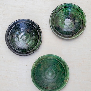 May include: Three ceramic bowls with a dark green glaze. The bowls are round with a slightly raised rim. The glaze has variations in colour, ranging from dark green to lighter shades. The bowls are arranged on a light-coloured surface.