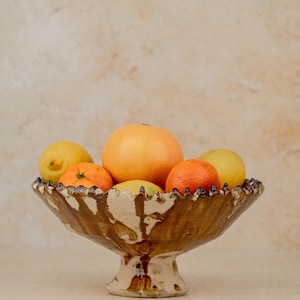May include: A decorative ceramic fruit bowl filled with citrus fruits. The bowl is brown with a cream interior and a scalloped rim. The fruits include oranges, grapefruits, and lemons.