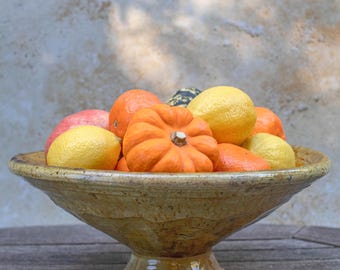 Puede incluir: Un cuenco de cerámica rústico sobre pedestal, lleno de una variedad de productos frescos. El cuenco es de un tono cálido y terroso, y contiene naranjas, limones, una pequeña calabaza y otras frutas. El arreglo está sobre una superficie de madera.