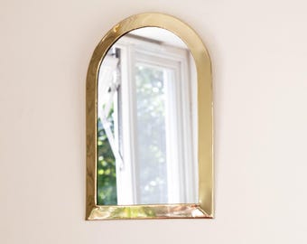 Può includere: Uno specchio dorato ad arco con un bordo bisellato. Lo specchio riflette una finestra con vista sugli alberi.