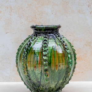 May include: A handcrafted, green ceramic vase with a textured, ridged design. The vase has a glossy finish with streaks of orange and yellow. The decorative vase has a round body and a narrow neck. The vase is approximately 25 cm tall.