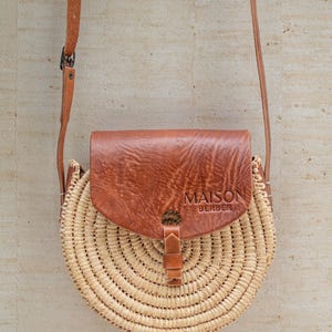 May include: A circular woven straw bag with a brown leather flap and strap. The flap is embossed with "MAISON BERBER". The bag has a buckle closure and a long, adjustable strap.