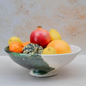 May include: A ceramic fruit bowl, half glazed in green and half in white, filled with a variety of fresh fruits. The bowl contains a red pomegranate, oranges, lemons, and a small decorative gourd.