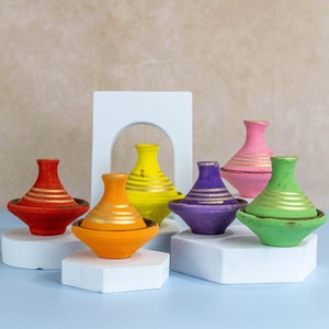 Puede incluir: Seis tagines de cerámica de colores con detalles dorados, dispuestos en plataformas blancas. Las tagines son rojas, naranjas, amarillas, moradas, verdes y rosas.
