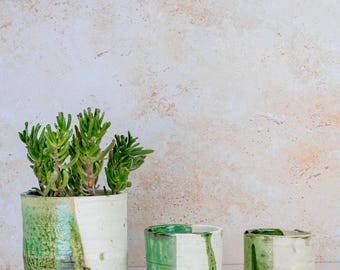 Puede incluir: Tres macetas de cerámica hechas a mano en fila, cada una con un diseño único. La maceta más grande contiene una planta suculenta verde. Las macetas tienen una base blanca con detalles de esmalte verde, creando un aspecto rústico y artístico. Adecuado para uso en interiores o exteriores.