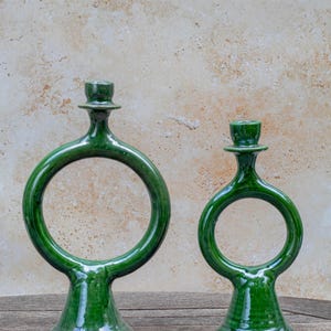 May include: Two green ceramic candle holders with a circular design. The candle holders have a glossy finish and are placed on a wooden surface. The background is a textured, neutral colour.