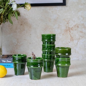 May include: A set of six green ceramic cups with a textured finish. The cups are stacked in a pyramid shape, with the largest cup at the bottom and the smallest cup at the top.