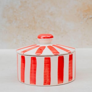 Puede incluir: Un tarro redondo de cerámica con tapa, con rayas verticales rojas sobre fondo blanco. La tapa tiene un patrón de rayas rojas y blancas a juego y un pequeño pomo redondo rojo. Ideal para almacenamiento y decoración.