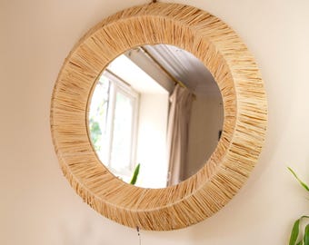 May include: A round mirror with a natural woven frame made of straw-like material. The mirror is hanging on a light beige wall.