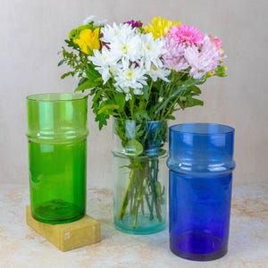 May include: Three glass vases, one green, one blue, and one clear, with a bouquet of white, pink, and yellow flowers.