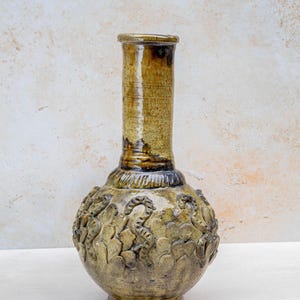 May include: A tall, handcrafted ceramic vase with a textured, cylindrical neck and a rounded body. The vase features a detailed, raised relief pattern around the body and is finished in shades of brown and olive green.