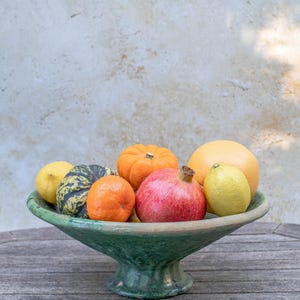 May include: A green ceramic pedestal bowl filled with a variety of fresh fruits. The bowl contains a small orange pumpkin, a pomegranate, a grapefruit, a tangerine, a lemon, and a small squash. The bowl sits on a wooden table.