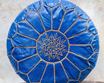 May include: A round, blue leather pouf with a geometric design. The pouf features a central starburst pattern and is stitched with gold-colored thread. The overall design is a combination of geometric shapes and intricate details.