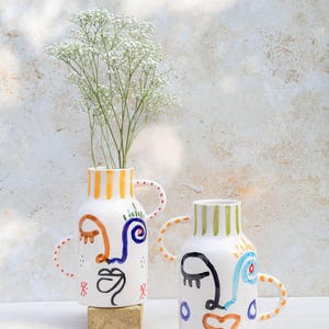 Puede incluir: Dos jarrones de cerámica blanca con diseños de caras abstractas. Un jarrón contiene flores blancas. Los jarrones tienen detalles coloridos, incluyendo rayas y asas naranjas, azules y verdes. Los jarrones están sobre una superficie blanca.