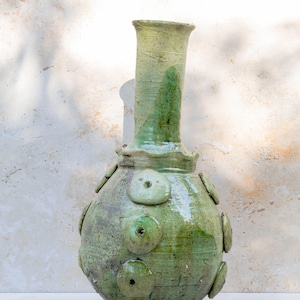 Könnte beinhalten: Eine handgefertigte grüne Keramikvase mit bauchigem Körper und hohem, zylindrischem Hals. Die Vase hat kreisförmige, erhabene Verzierungen und eine strukturierte Oberfläche. Die Farbe ist eine Mischung aus hellen und dunklen Grüntönen mit glänzendem Finish.
