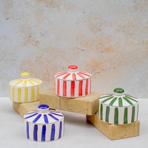 May include: Four ceramic lidded jars with vertical stripes in yellow, red, green, and blue. Each jar has a matching coloured knob on the lid. The jars are arranged on a light-coloured surface with a textured background.