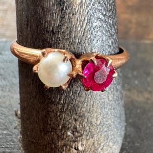 May include: A gold ring featuring two stones set side-by-side in a bypass style. One stone is a smooth, round white pearl, and the other is a faceted, vibrant pinkish-red gemstone. The metal band has a polished finish and a warm, golden hue.