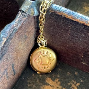 May include: A small, round, gold-toned metal locket pendant suspended from a delicate gold-toned chain. The front surface of the locket features an engraved geometric pattern with a central square motif and decorative floral-like accents.