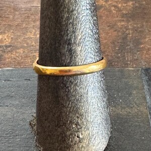 May include: A gold ring displayed on a wooden ring holder. The ring has a simple, rounded band and a bright, polished finish. The ring is a classic piece of jewelry.