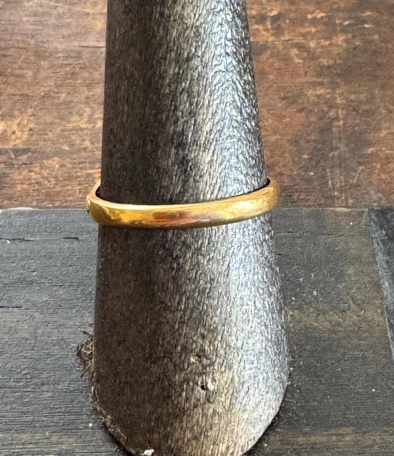 May include: A gold ring displayed on a wooden ring holder. The ring has a simple, rounded band and a bright, polished finish. The ring is a classic piece of jewelry.