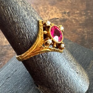 May include: A gold ring featuring an oval, bright pink gemstone as the centerpiece, surrounded by smaller white stones. The ring has an ornate, filigree design with a detailed band. The ring is displayed on a dark wooden ring holder.