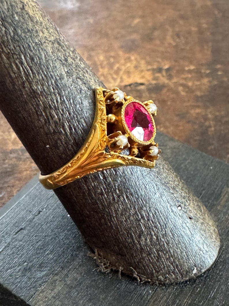 May include: A gold ring featuring an oval, bright pink gemstone as the centerpiece, surrounded by smaller white stones. The ring has an ornate, filigree design with a detailed band. The ring is displayed on a dark wooden ring holder.