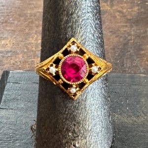 May include: A gold ring with a large, round, red gemstone at the center, surrounded by smaller white stones and black accents. The ring has an ornate, geometric design.