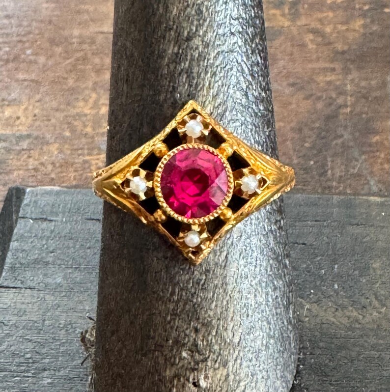 May include: A gold ring with a large, round, red gemstone at the center, surrounded by smaller white stones and black accents. The ring has an ornate, geometric design.