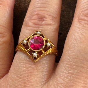 May include: A gold ring featuring a large, round, red gemstone at the center, surrounded by small pearls. The ring has an ornate, filigree design with a diamond-shaped setting. The ring is on a finger.
