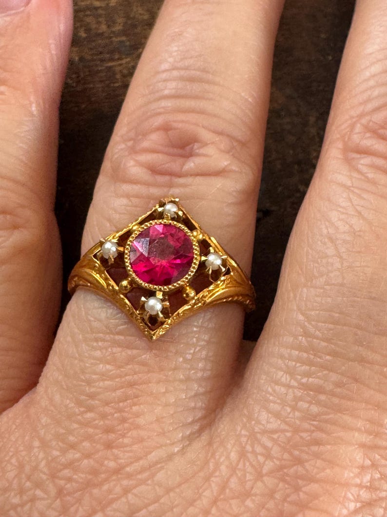 May include: A gold ring featuring a large, round, red gemstone at the center, surrounded by small pearls. The ring has an ornate, filigree design with a diamond-shaped setting. The ring is on a finger.
