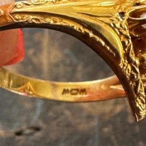 May include: Close-up of a gold ring with intricate filigree detailing. The ring has a decorative design on the top and the word "MOM" is engraved on the inside of the band.