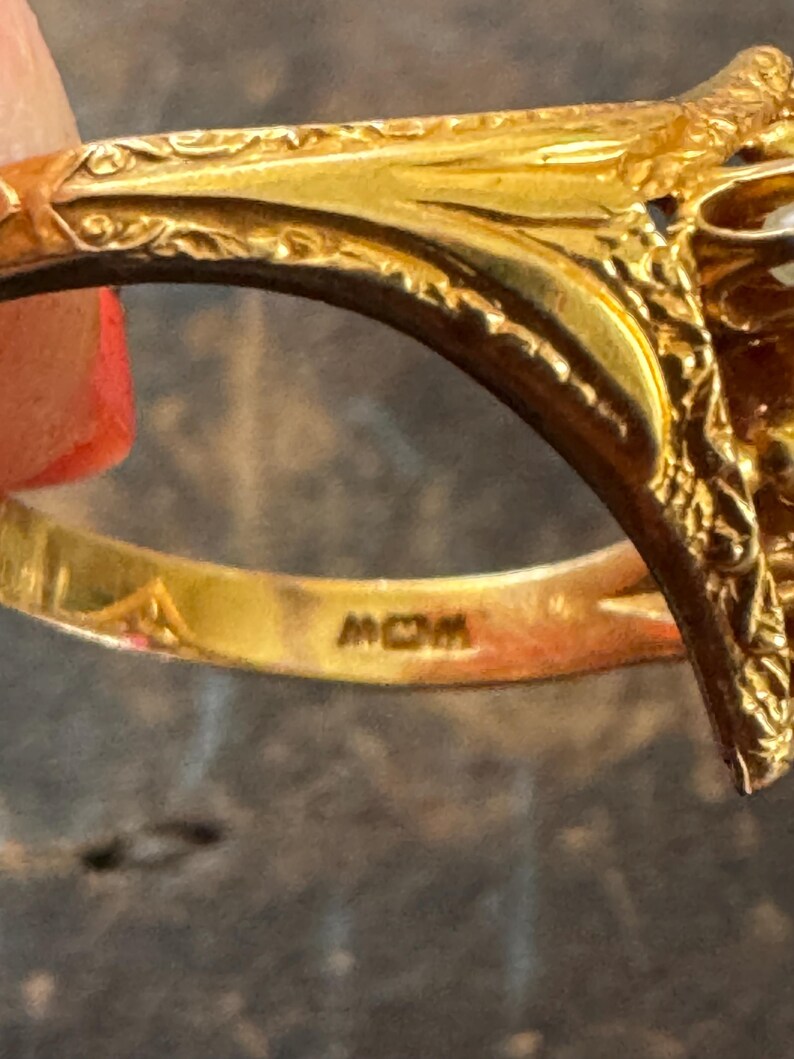 May include: Close-up of a gold ring with intricate filigree detailing. The ring has a decorative design on the top and the word "MOM" is engraved on the inside of the band.