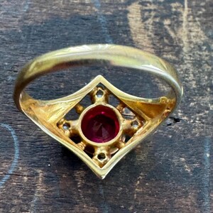 May include: A gold ring with a central, round, red gemstone. The gemstone is set within a diamond-shaped frame, accented with smaller, clear stones. The ring band is a simple, polished gold.
