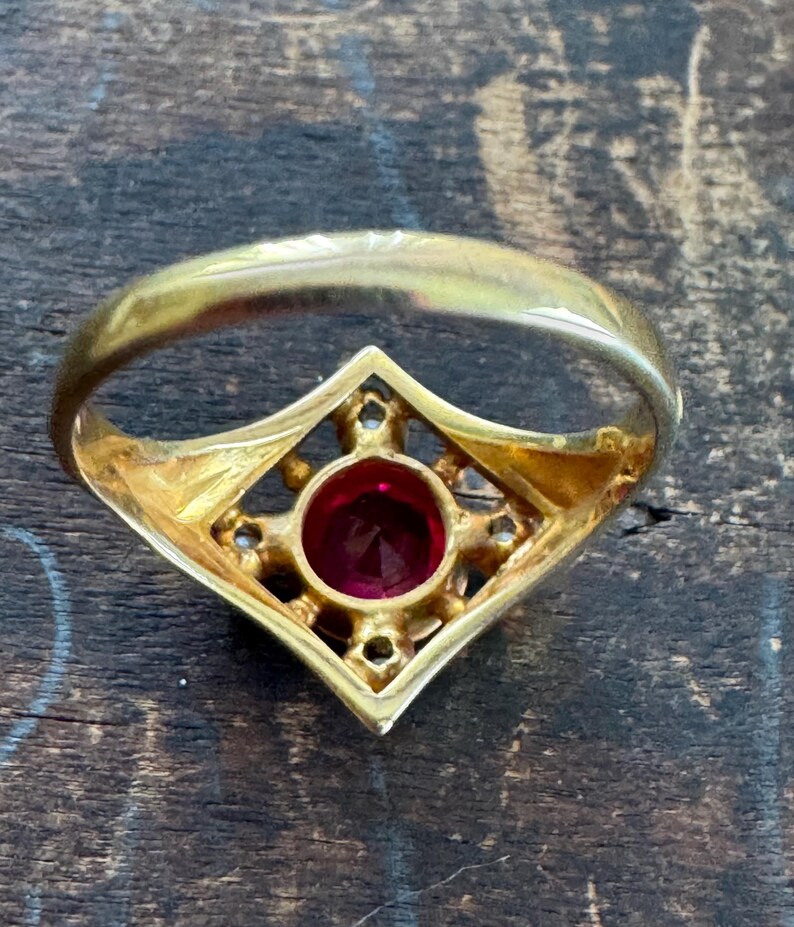May include: A gold ring with a central, round, red gemstone. The gemstone is set within a diamond-shaped frame, accented with smaller, clear stones. The ring band is a simple, polished gold.