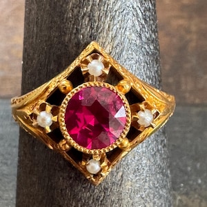 May include: A gold-colored ring featuring a large, round, ruby-red gemstone at the center. The ring has an ornate, filigree design with small, white pearl accents. The setting is a diamond shape.