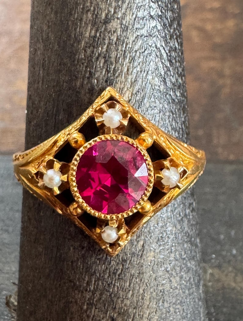 May include: A gold-colored ring featuring a large, round, ruby-red gemstone at the center. The ring has an ornate, filigree design with small, white pearl accents. The setting is a diamond shape.