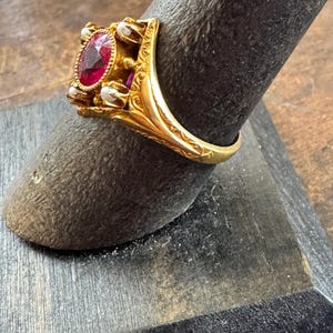 May include: A gold ring featuring an oval, deep red gemstone at the center, surrounded by small, white pearls. The ring has an ornate, detailed design with engraved patterns on the band and setting.