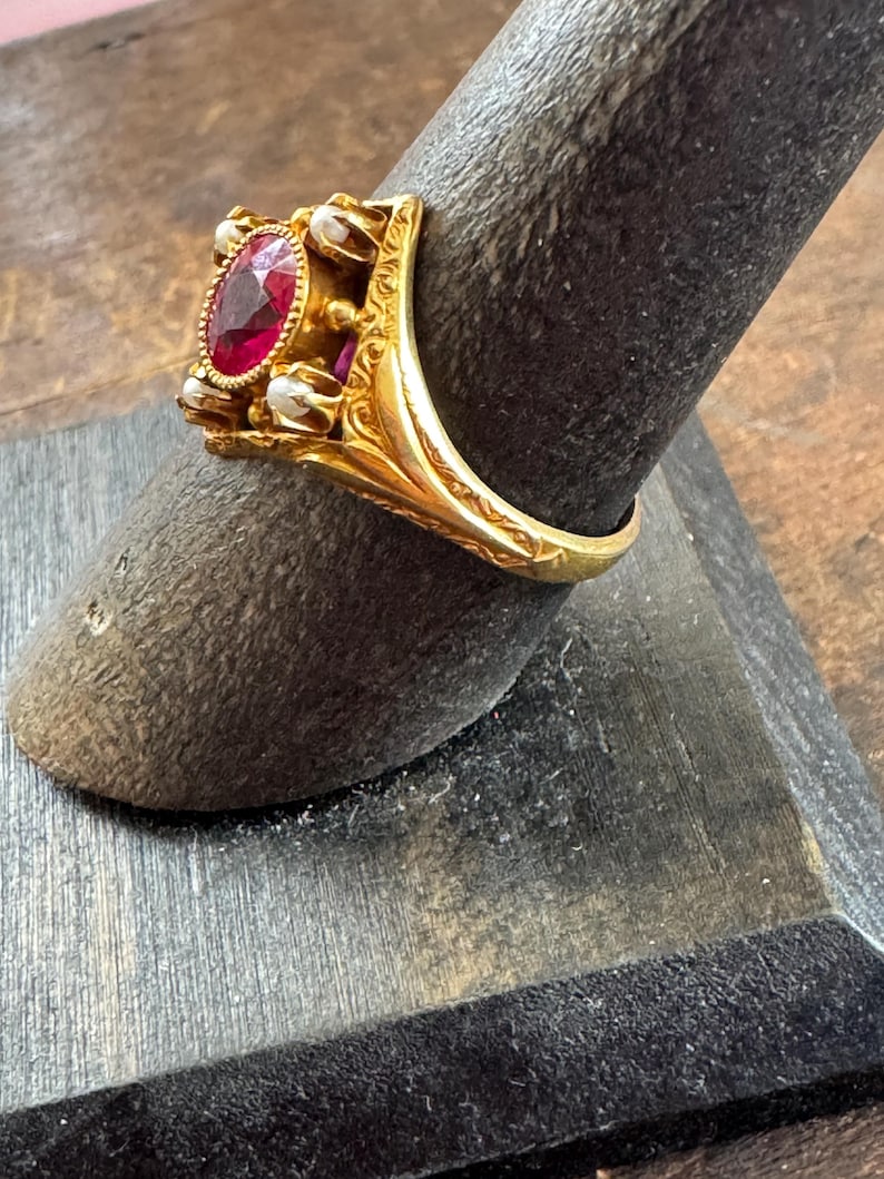 May include: A gold ring featuring an oval, deep red gemstone at the center, surrounded by small, white pearls. The ring has an ornate, detailed design with engraved patterns on the band and setting.