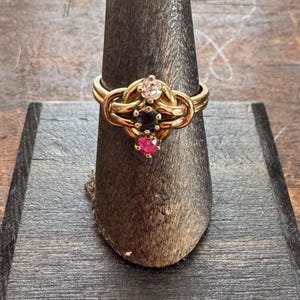 May include: A gold-toned ring with a knot design, featuring three gemstones: a clear, a dark blue, and a red stone. The ring is displayed on a wooden ring holder, showcasing its intricate details and craftsmanship.