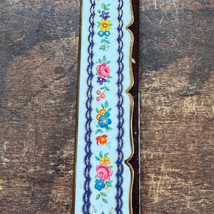 May include: A rectangular, vintage bookmark with a floral design. The bookmark features a light blue background with a central vertical band of colorful flowers, framed by a dark blue border and gold trim. The back is a dark brown color.