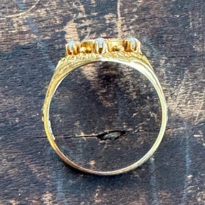 May include: A gold ring with a central red gemstone and surrounding smaller stones. The ring's band is detailed with etched patterns. The ring is set against a dark wooden surface.