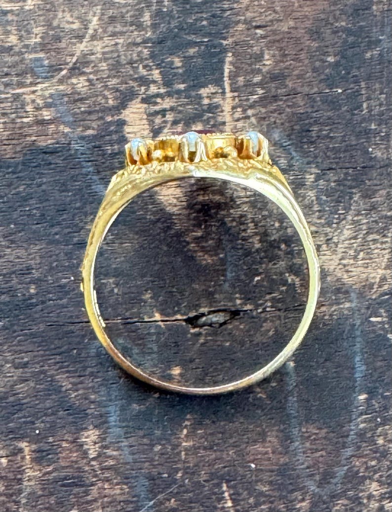 May include: A gold ring with a central red gemstone and surrounding smaller stones. The ring's band is detailed with etched patterns. The ring is set against a dark wooden surface.