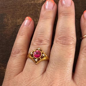 May include: A gold ring featuring a central, round, red gemstone set within a square frame. The frame is adorned with small, white pearls and intricate gold filigree detailing. The ring is on a hand with orange nail polish.