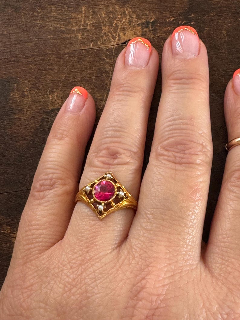 May include: A gold ring featuring a central, round, red gemstone set within a square frame. The frame is adorned with small, white pearls and intricate gold filigree detailing. The ring is on a hand with orange nail polish.