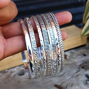 Pode incluir: Conjunto de seis pulseiras de prata com diferentes desenhos em relevo. As pulseiras estão empilhadas umas sobre as outras e têm um acabamento brilhante.