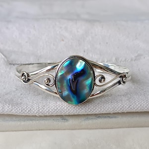 Puede incluir: Un brazalete de plata con una gran incrustación ovalada de concha de abulón. La concha tiene colores iridiscentes azules, verdes y blancos.